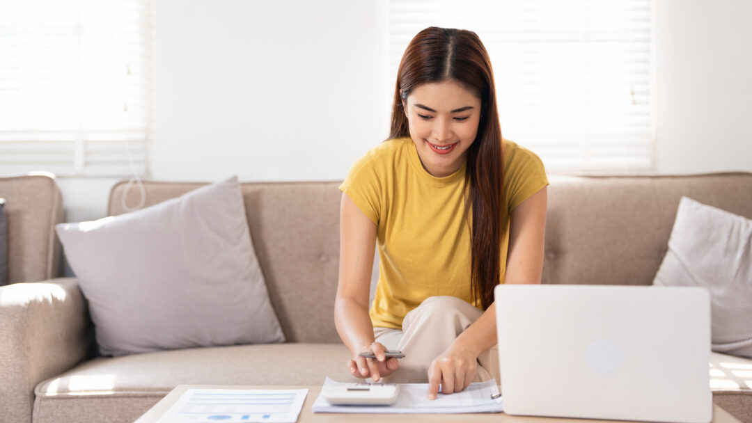 Woman using a calculator and laptop to review monthly budget at home.