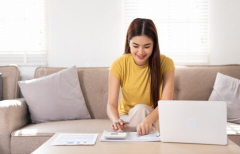Woman using a calculator and laptop to review monthly budget at home.