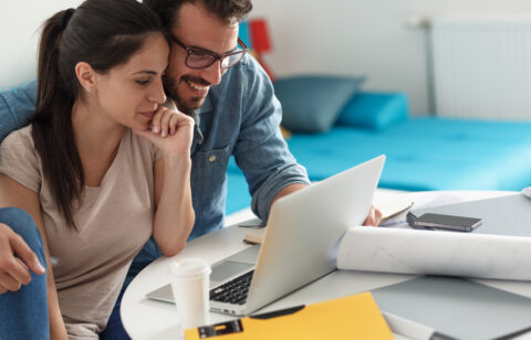 A couple sits together at a table reviewing financial documents and plans on a laptop.