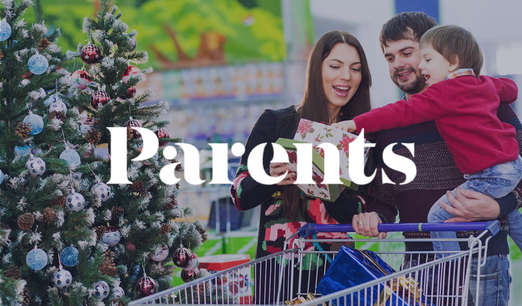 A smiling family shopping for holiday gifts, standing by a decorated Christmas tree with a cart full of presents. The Parents logo is overlayed.