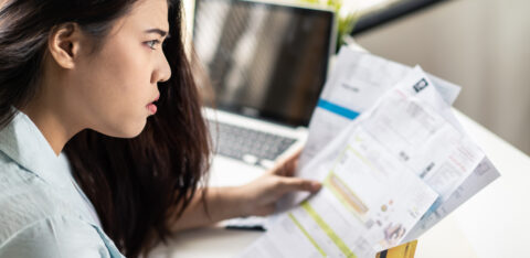 A woman sits at her desk holding several bills and a credit card, looking worried as she reviews her finances in front of a laptop.