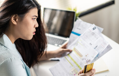 A woman sits at her desk holding several bills and a credit card, looking worried as she reviews her finances in front of a laptop.