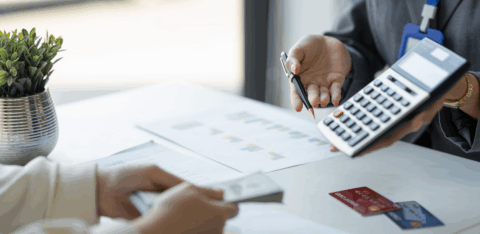 A person holds a calculator and pen while discussing paperwork with someone holding cash at a desk.