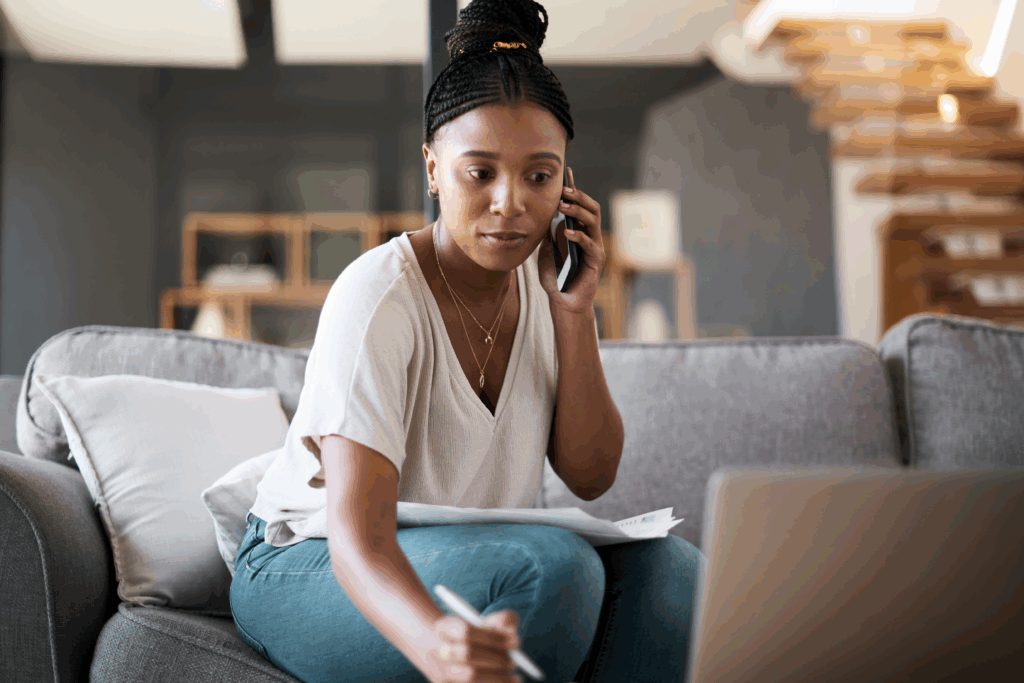A woman sitting on a couch talks on the phone while reviewing papers and taking notes on her laptop.