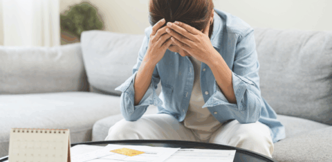 A woman sits at her coffee table with bills and a calculator, holding her head in stress while reviewing her finances.