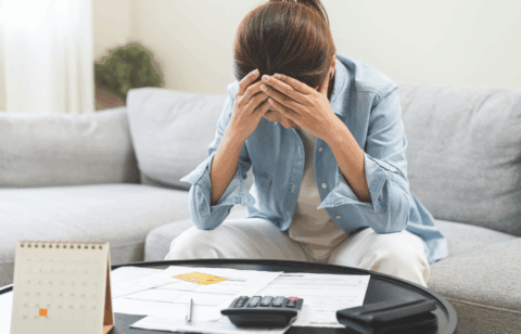 A woman sits at her coffee table with bills and a calculator, holding her head in stress while reviewing her finances.