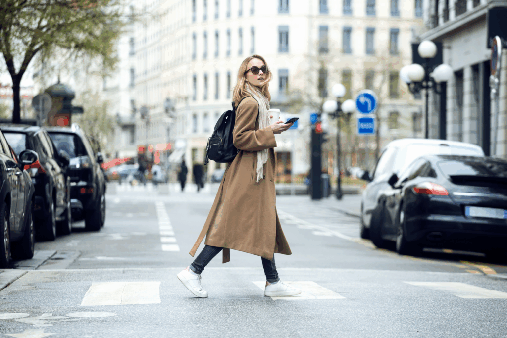 A woman wearing a long coat and backpack crosses a city street while holding a smartphone and a coffee.