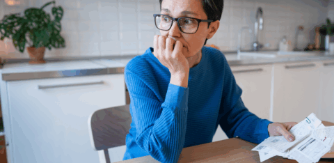 Person sitting at a kitchen table holding bills and looking off to the side with a thoughtful expression.