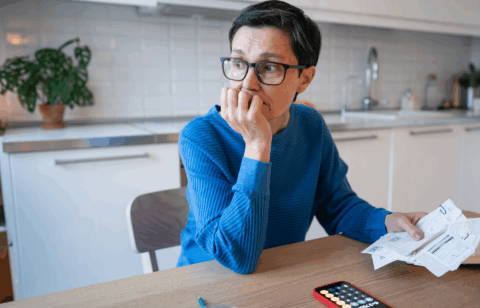 Person sitting at a kitchen table holding bills and looking off to the side with a thoughtful expression.