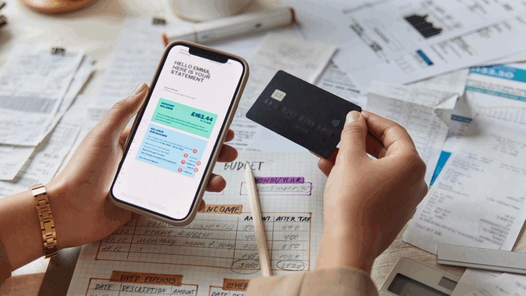 Hands hold a smartphone showing a bank statement and a credit card above a table covered with bills, receipts, and a handwritten budget.