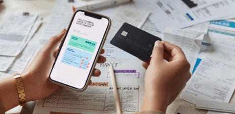 Hands hold a smartphone showing a bank statement and a credit card above a table covered with bills, receipts, and a handwritten budget.