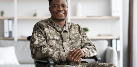 Veteran in camouflage uniform seated in a wheelchair, smiling at the camera.