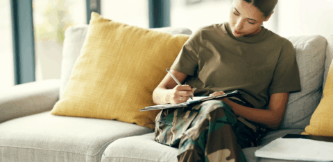 A service member sits on a couch and writes on a clipboard while reviewing paperwork.