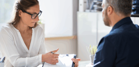 A woman wearing glasses points to a document on a clipboard while speaking with a man seated across a desk.