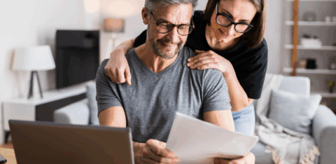 A middle-aged couple reviews paperwork together at a table while looking at a laptop in their living room.