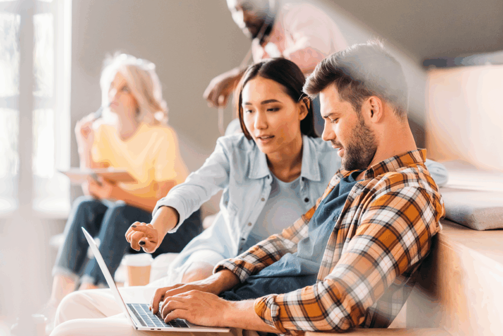 A woman points at a laptop screen while working with a man during a group study or workshop.