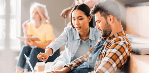 A woman points at a laptop screen while working with a man during a group study or workshop.