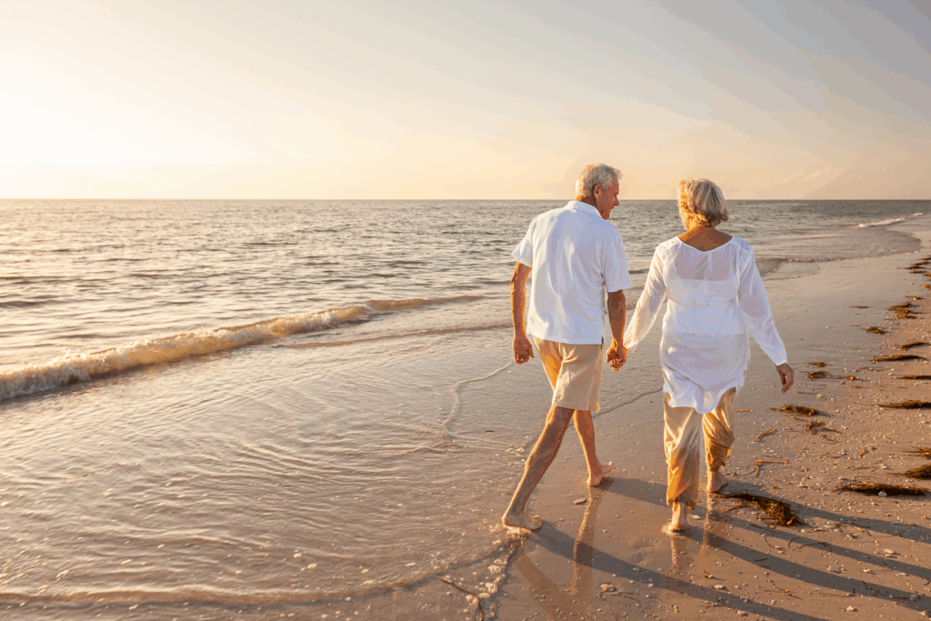 An older couple walks hand in hand along the beach at sunset.