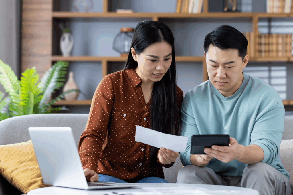 A couple sits on a couch reviewing a bill together while using a calculator and a laptop.