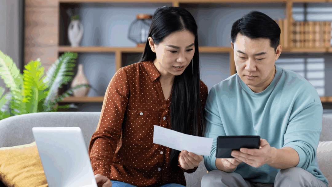 A couple sits on a couch reviewing a bill together while using a calculator and a laptop.