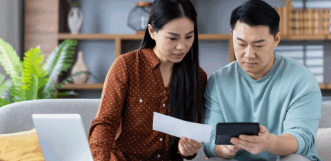 A couple sits on a couch reviewing a bill together while using a calculator and a laptop.