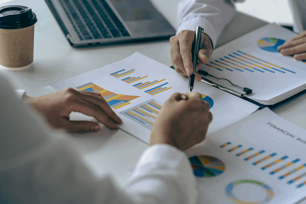 Two people reviewing printed charts and graphs at a desk, pointing at data with a pen near a laptop.