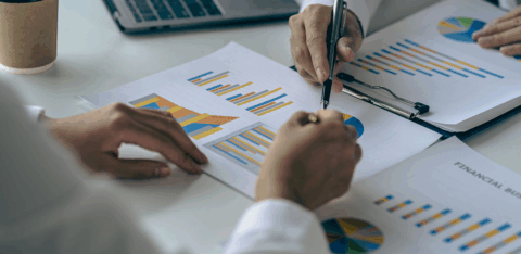 Two people reviewing printed charts and graphs at a desk, pointing at data with a pen near a laptop.