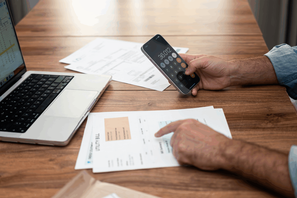 Person reviewing household bills at a table while using a calculator app on their phone next to a laptop.