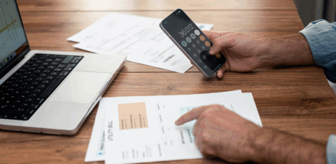 Person reviewing household bills at a table while using a calculator app on their phone next to a laptop.