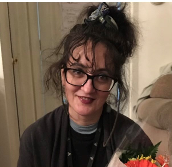 A woman with glasses and dark hair pulled up smiles while holding a bouquet of flowers indoors.