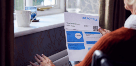 A person in a cozy room reads a printed energy bill while resting a hand on a radiator.
