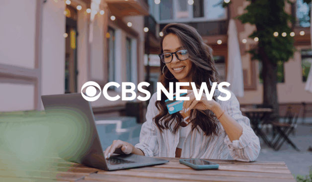 A person sitting outdoors uses a laptop while holding a credit card, with a CBS News logo overlaid on the image.