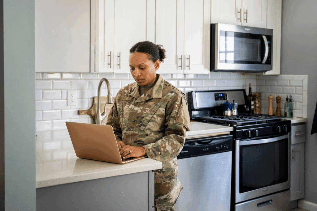 A woman in a military uniform stands at a kitchen counter using a laptop in her home.
