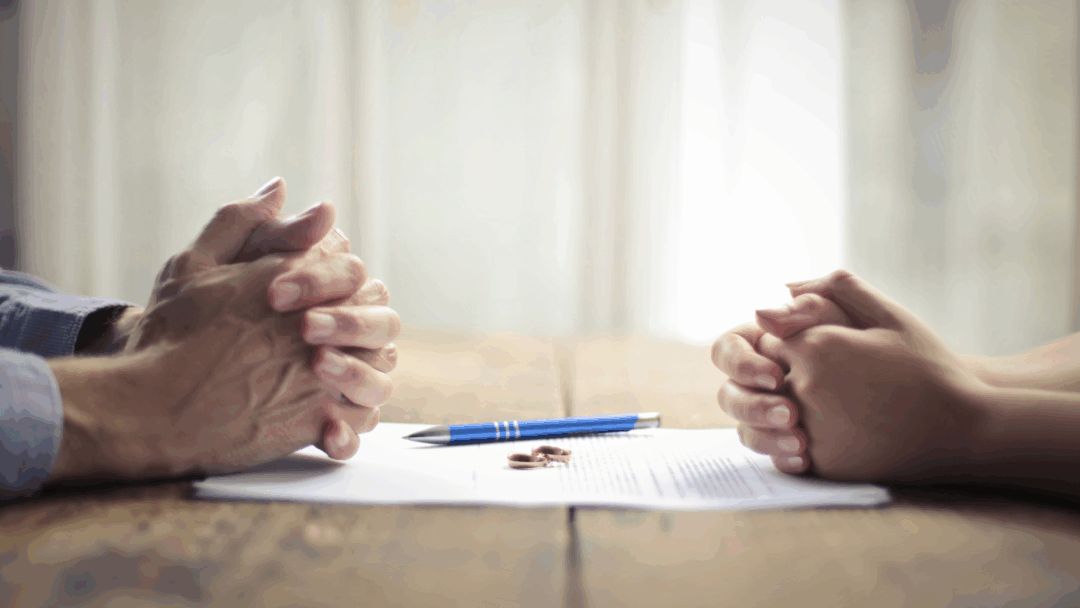 Two people sit across from each other at a table with clasped hands, a document, a pen, and wedding rings placed between them.