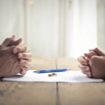 Two people sit across from each other at a table with clasped hands, a document, a pen, and wedding rings placed between them.