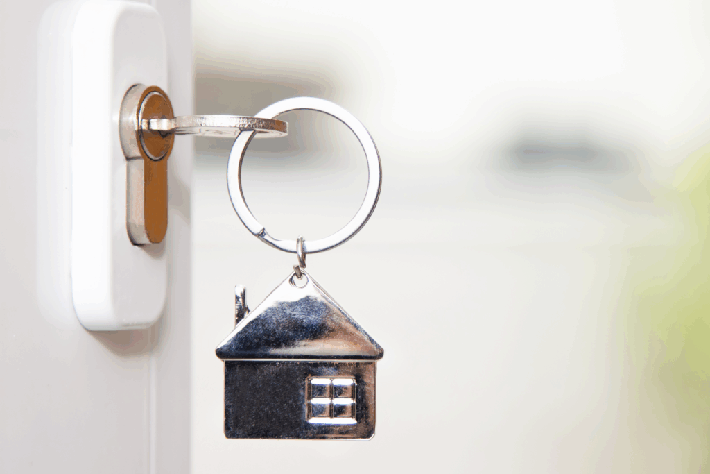 Close-up of a door key with a small metal house keychain attached, shown against a soft background.