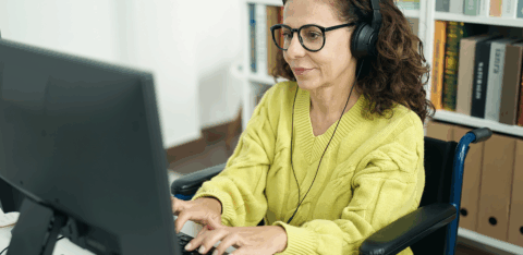 Woman wearing glasses and headphones uses a desktop computer while seated in a wheelchair in a home office.