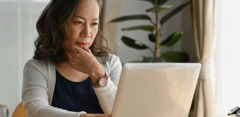 Older woman sitting at a table using a laptop, appearing focused while reviewing information at home.