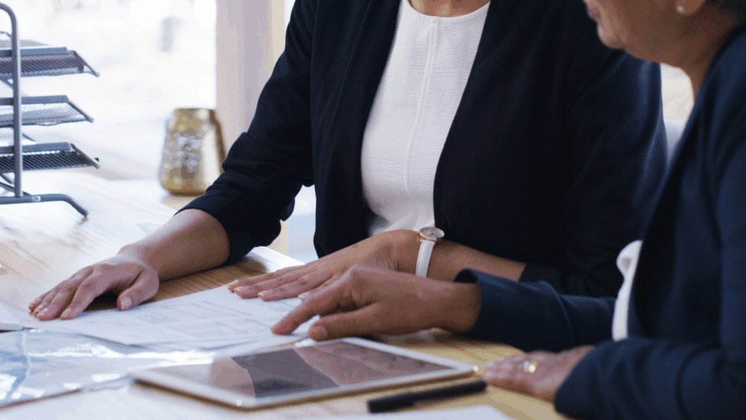 Two people reviewing paperwork together at a desk, with documents and a tablet laid out in front of them.