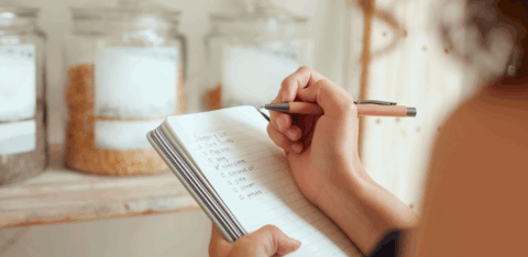 Person writing a grocery shopping list in a notebook with pantry jars in the background.