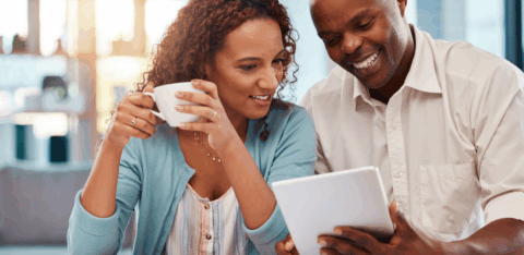 Couple sitting at a table reviewing financial documents on a tablet while drinking coffee at home.