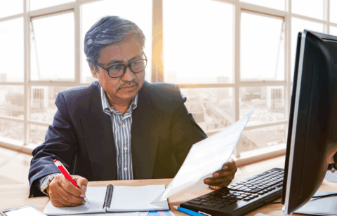 Professional reviewing documents while taking notes at a desk, with a desktop computer and papers nearby.