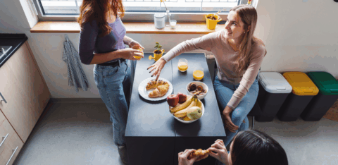 Three young adults sharing breakfast at a kitchen island with fruit, pastries, and drinks.