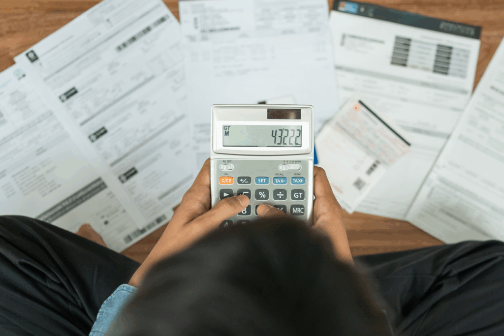 Person using a calculator while reviewing bills and financial documents spread across a desk.