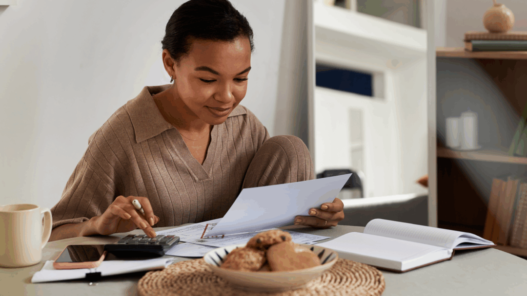 Woman sitting at a table reviewing bills and paperwork while using a calculator at home.