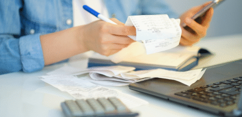 Person reviewing receipts while holding a phone, with a calculator and laptop on the desk.