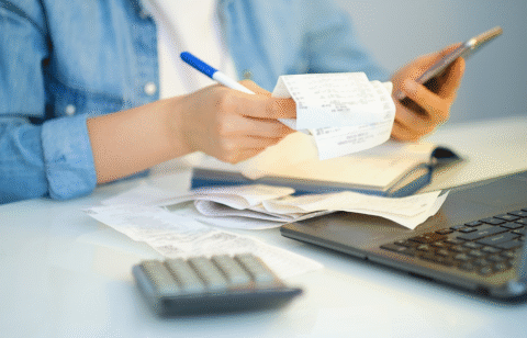 Person reviewing receipts while holding a phone, with a calculator and laptop on the desk.