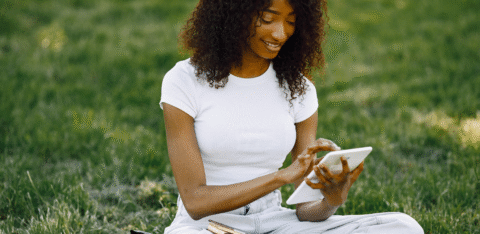 Woman sitting on grass using a tablet, with notebooks and books beside her.