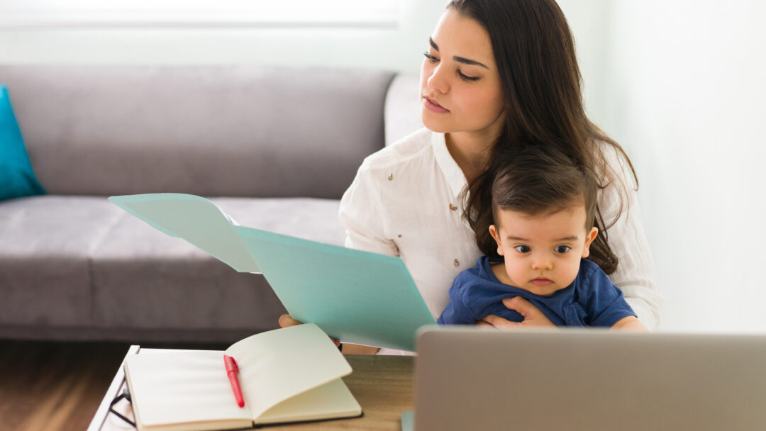 Woman holding a baby while reviewing paperwork at a desk with a laptop and open notebook.