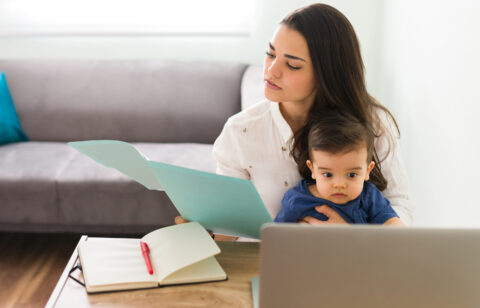 Woman holding a baby while reviewing paperwork at a desk with a laptop and open notebook.